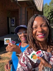Two women proudly display stickers outside a building, smiling and engaging with passersby.