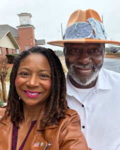 A man and woman in hats stand together in front of a church, smiling and enjoying the sunny day.